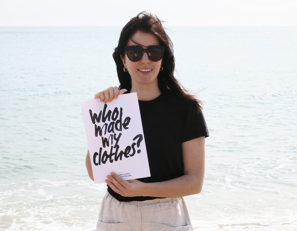 Besma standing by the sea holding up a sign saying ‘Who Made My Clothes?’