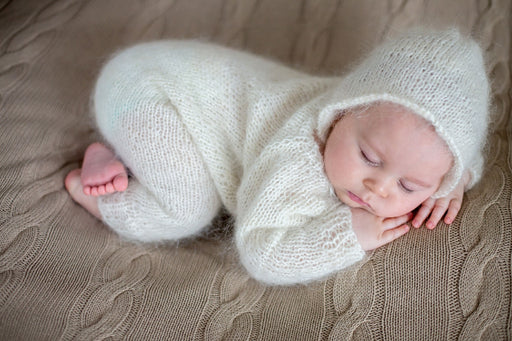 a gorgeous sleeping baby wearing a soft knitted all in one with hood lying on a cable knit blanket