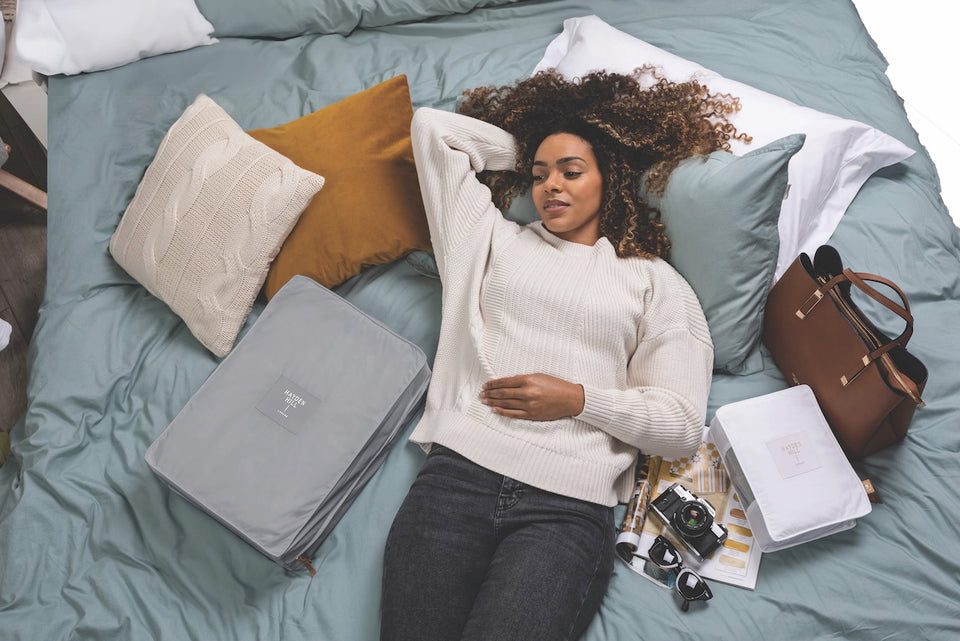 a woman lying on her bed surrounded by cushions, a purse and a two storage bags, one in gray and one in white