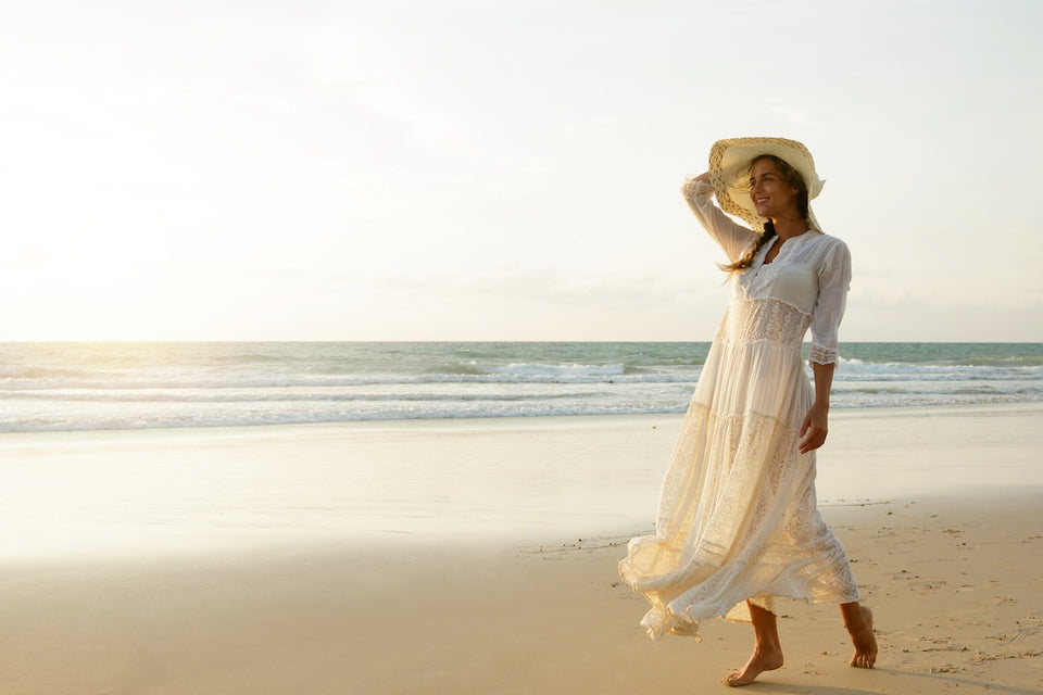 a woman enjoying a walk along the beach wearing a flowing white dress and straw hat