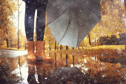 a woman, on a Fall day, standing in a puddle, wearing rain boots and holding an umbrella