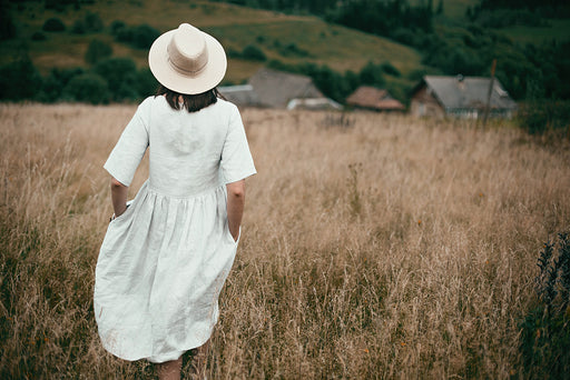 a woman wearing a linen dress and hat standing in a field.