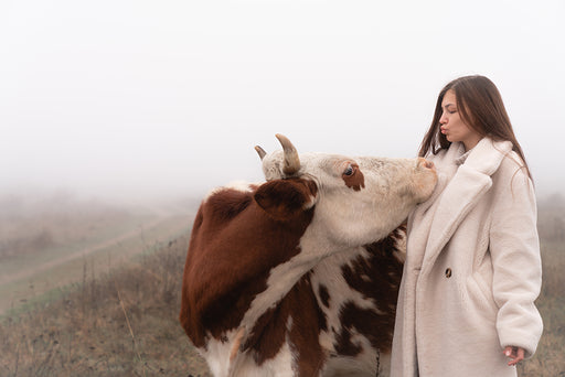 a woman wearing a thick cream coat blows a kiss to a cow