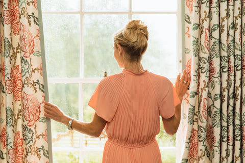 a woman wearing a peach colored silk dress looking out of the window through parted curtains