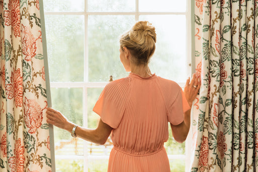 a woman wearing a peach colored silk dress looking out of the window through parted curtains