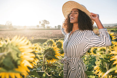 a laughing woman standing in a field of sunflowers wearing a stripy dress and sunhat