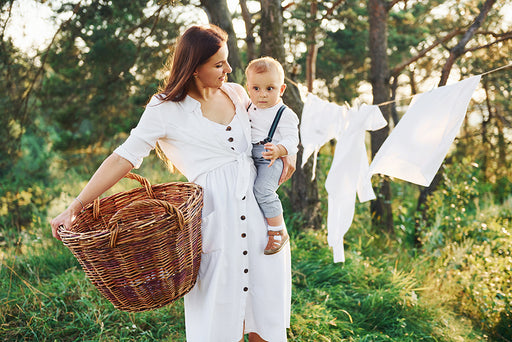 a mother outdoors drying her washing on the line while holding her child and a wicker laundry basket