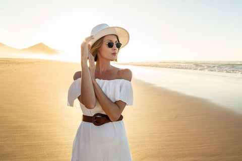 a woman on the beach wearing a white off the shoulder dress, brown belt, hat and sunglasses