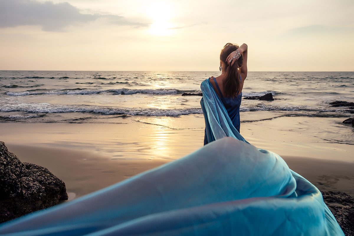a woman with a beautiful swathe of blue silk standing by the ocean at sunset