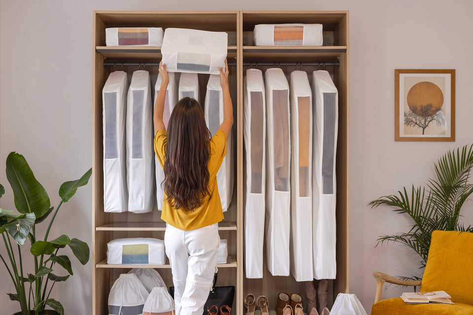 A woman putting an extra large Hayden Hill storage cube into an organized closet which is filled with hanging bags for clothes, short hanging garment bags, good-sized packing cubes, and multiple dust bags for purses, handbags or shoes.