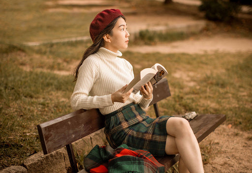 Mai sitting on a park bench in a tartan skirt, roll neck and beret