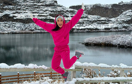 Riquel in a fuchsia pink hoodie and matching pants, jumping for joy in the mountains @R.Organize