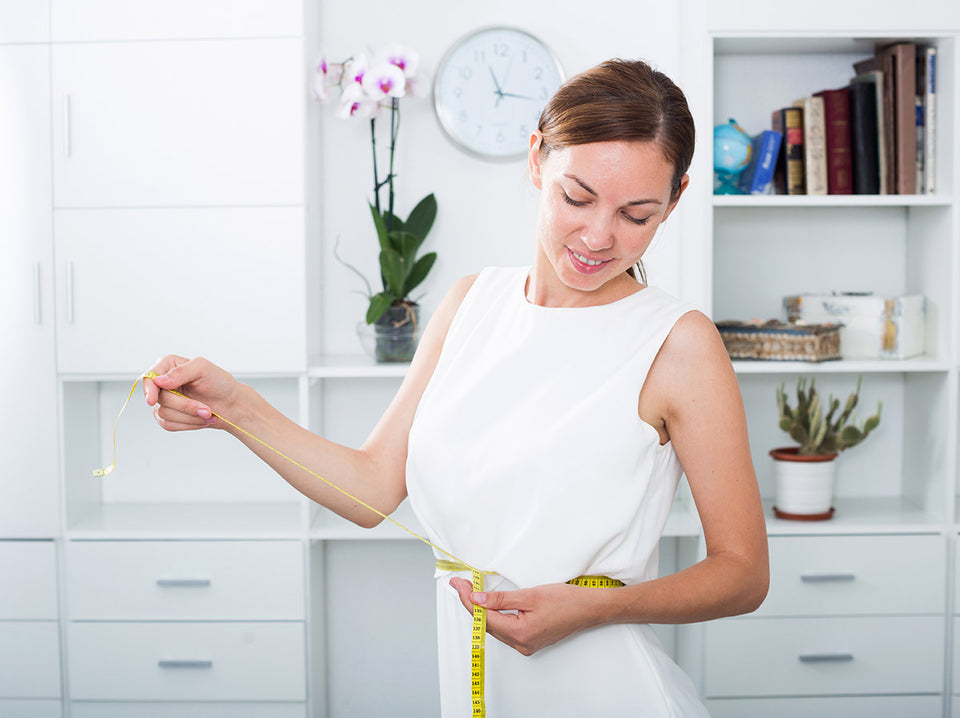 a woman at home dressed in white, measuring her waist