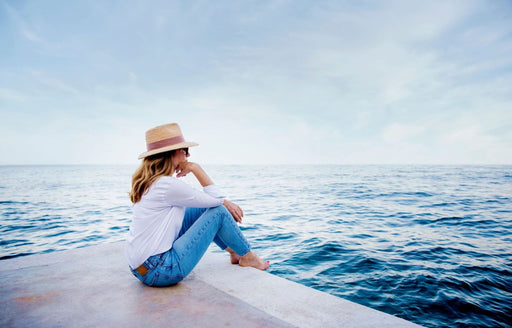 a woman in a white shirt denim jeans straw hat and sunglasses looking out to sea