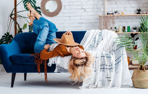 a woman in boyfriend jeans lying upside down on her sofa holding her hat