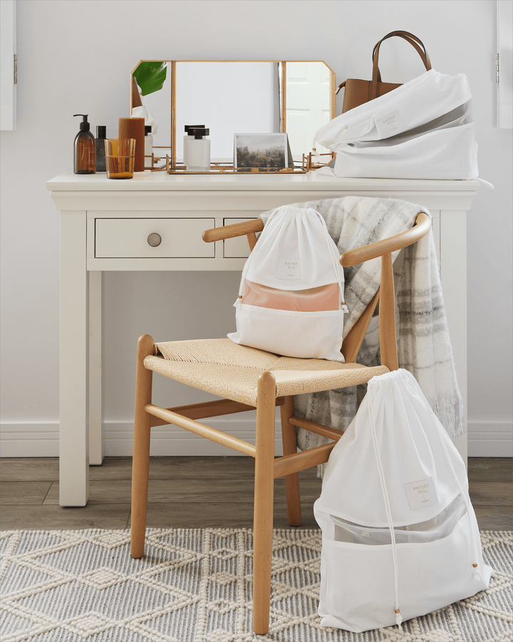 a medium dust bag with a purse inside it sitting on dressing table next to a small dust bag on a chair and a large dust bag beside the chair