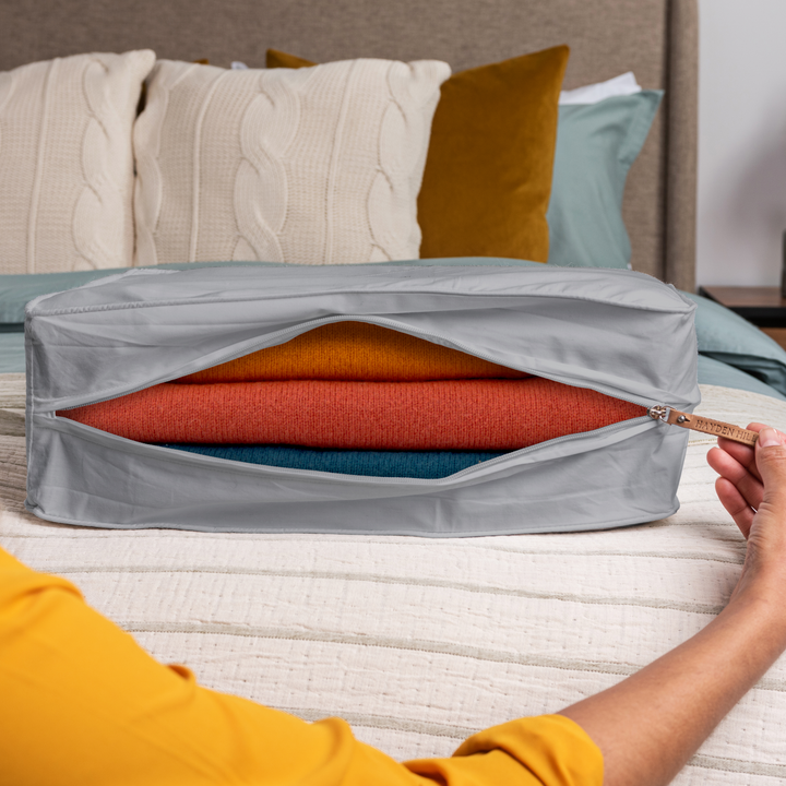 a grey extra large storage bag sitting on a bed being unzipped with a view of several woollen sweaters inside