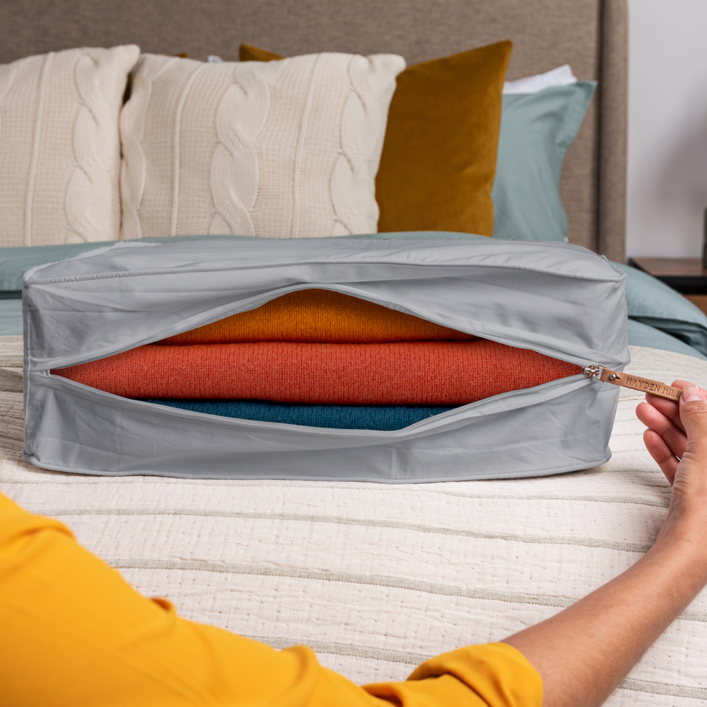 a grey extra large storage bag sitting on a bed being unzipped with a view of several woollen sweaters inside