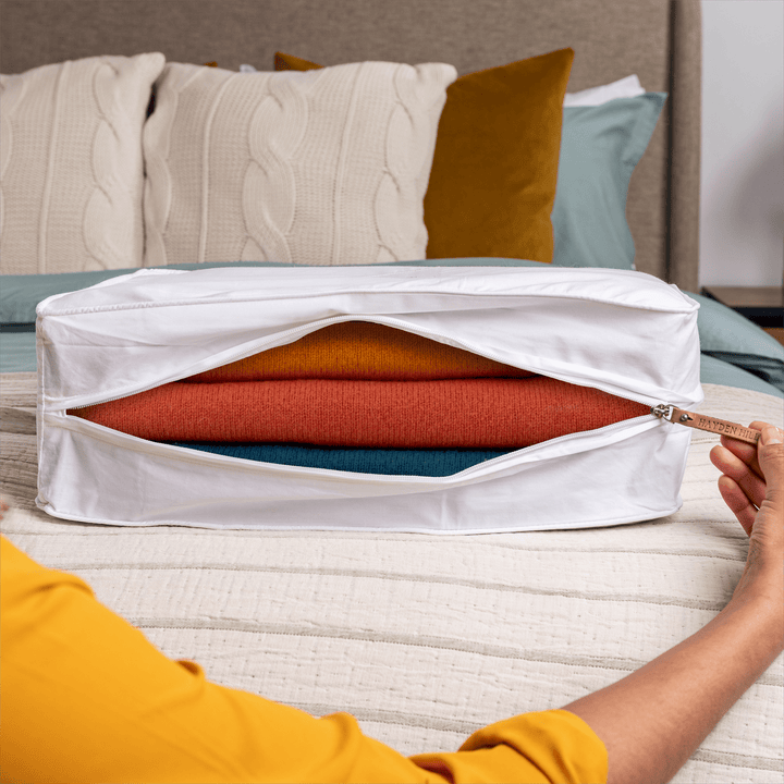 an extra large storage bag sitting on a bed being unzipped with a view of several woollen sweaters inside