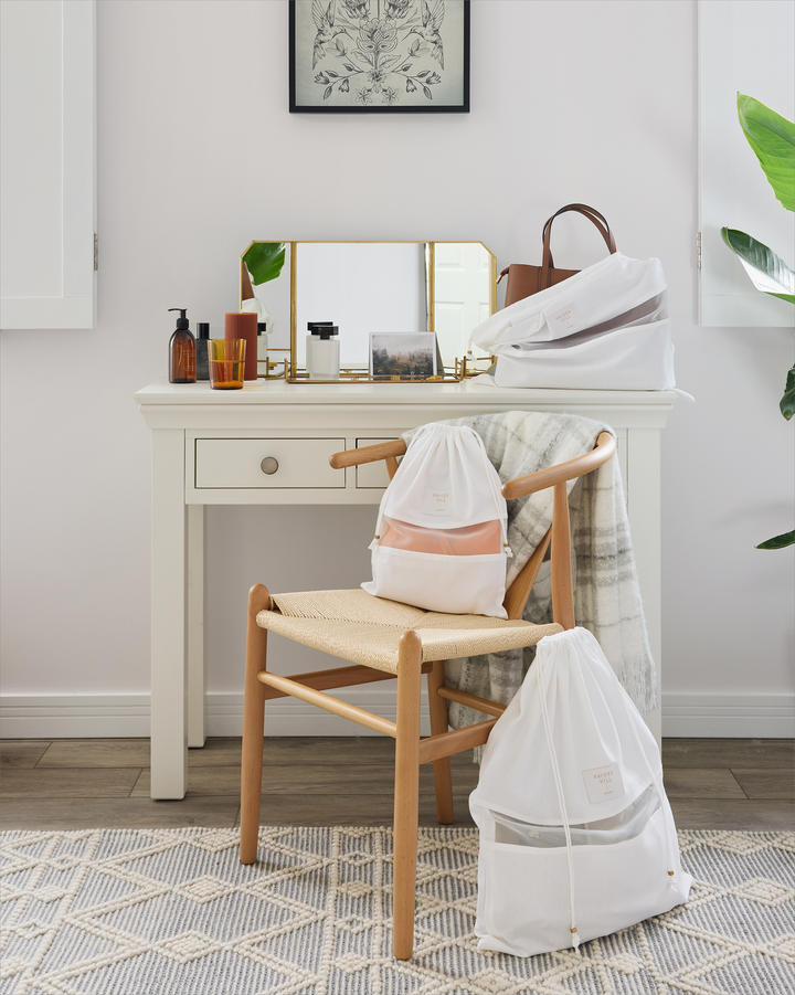 a medium dust bag with a purse inside it sitting on dressing table next to a small dust bag on a chair and a large dust bag beside the chair