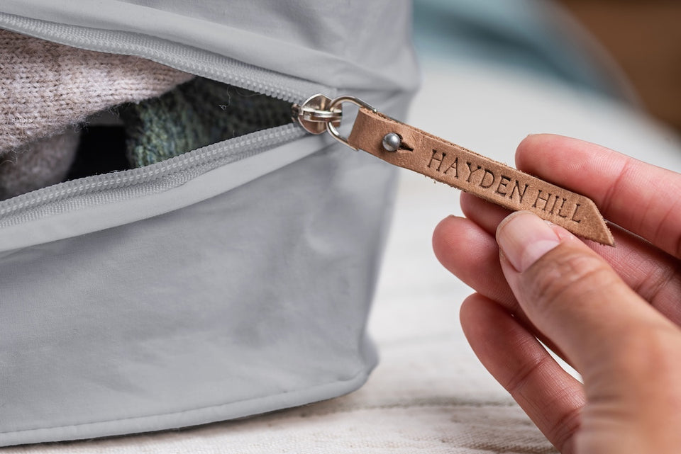 a close up of a hand showing the removable leather zipper pull on a gray Hayden Hill storage bag, filled with knitted sweaters